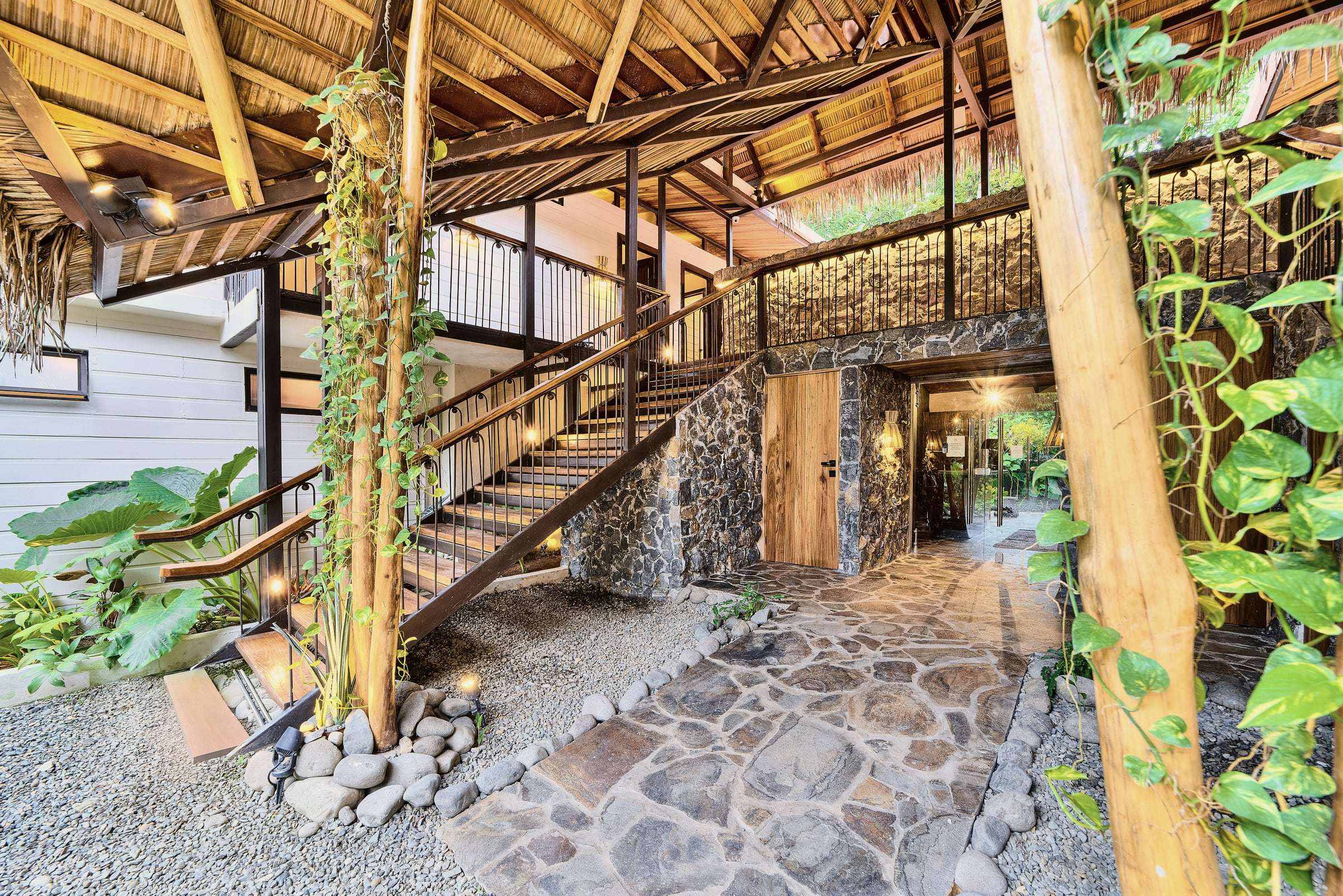 Atrium under the palm thatch: vine-wrapped wooden posts, an open staircase, stone walls and a flagstone path