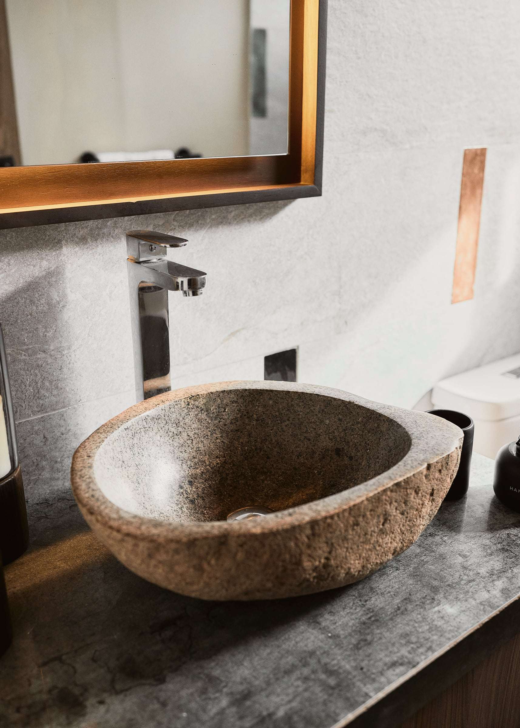 River-stone vessel sink with a chrome faucet on a dark stone counter, below a backlit wood-framed mirror