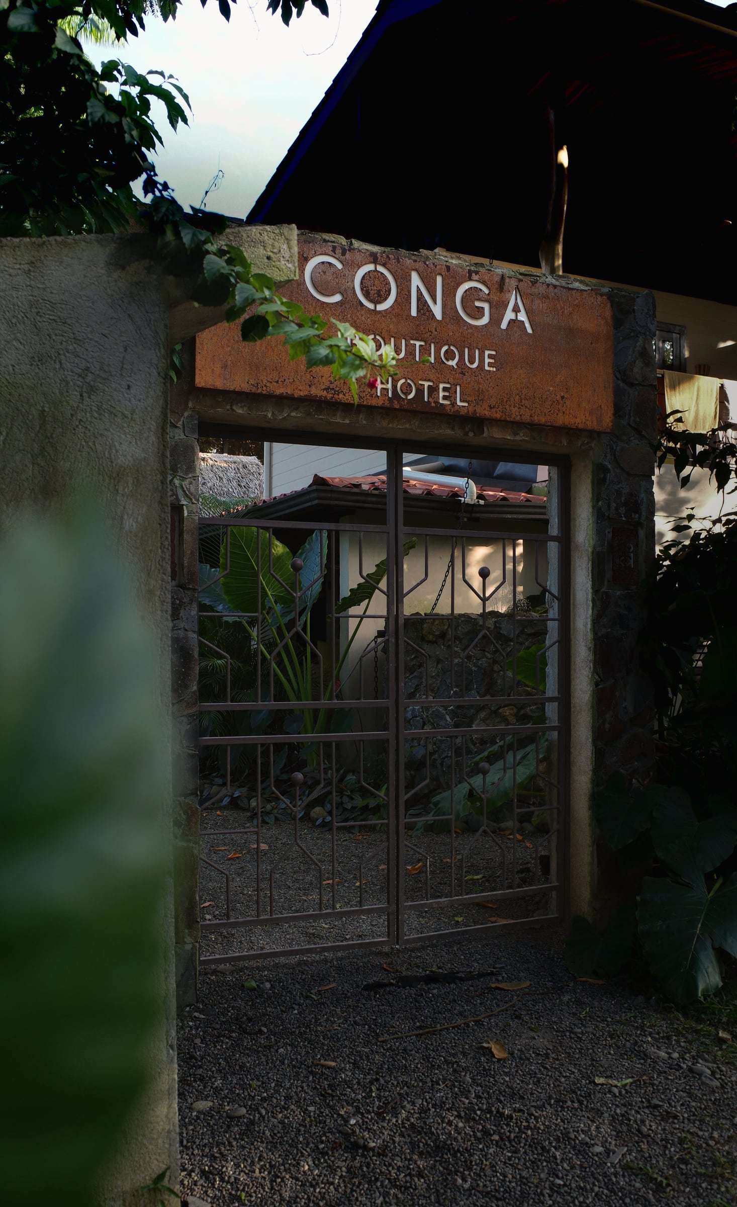 Iron street gate beneath the rusted-steel Conga Boutique Hotel sign, with the garden visible through the bars