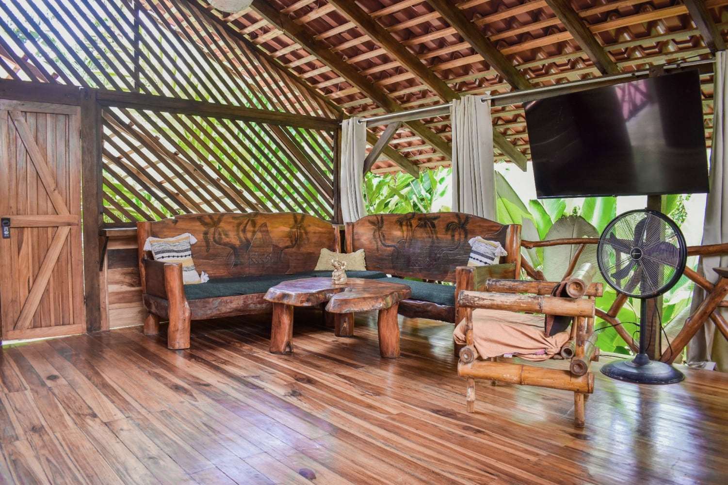 Garden Home open-air living room with carved wooden sofas, live-edge coffee tables and a wall-mounted TV