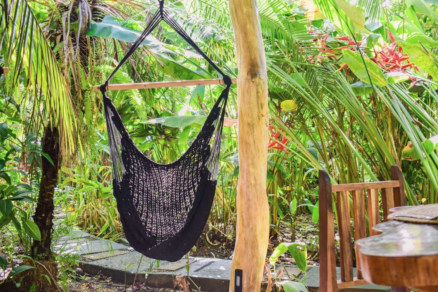 Black macramé hanging chair on a wooden post in the garden, among palms and red heliconias