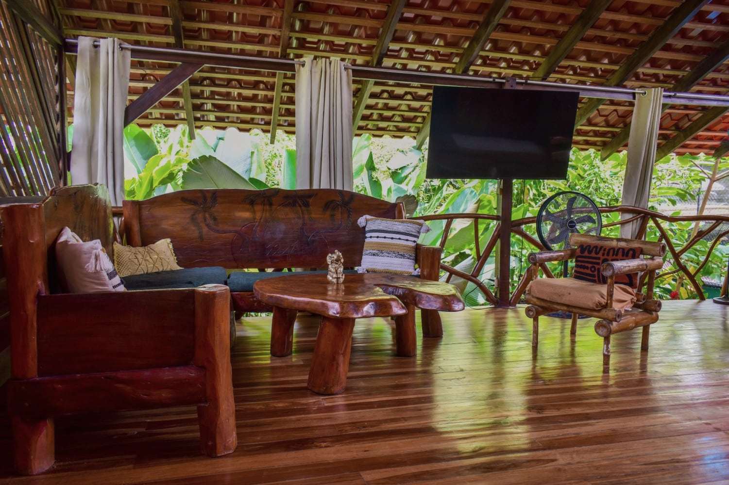 Garden Home: open-air living room under a tiled roof, with carved wooden sofas, live-edge tables and banana plants