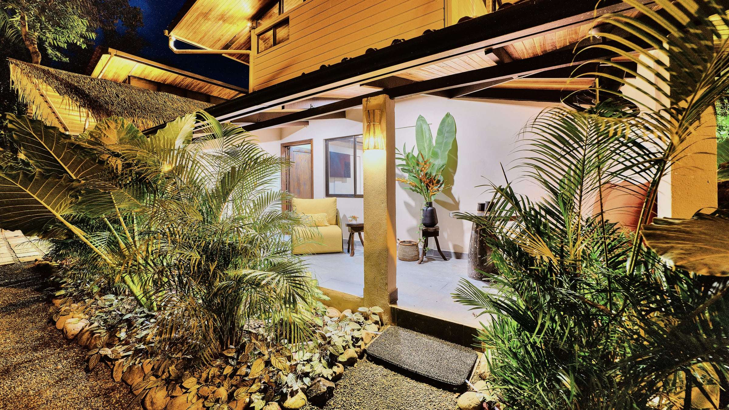 Ground-floor terrace at night with a yellow armchair, tropical flowers and palms lit from below