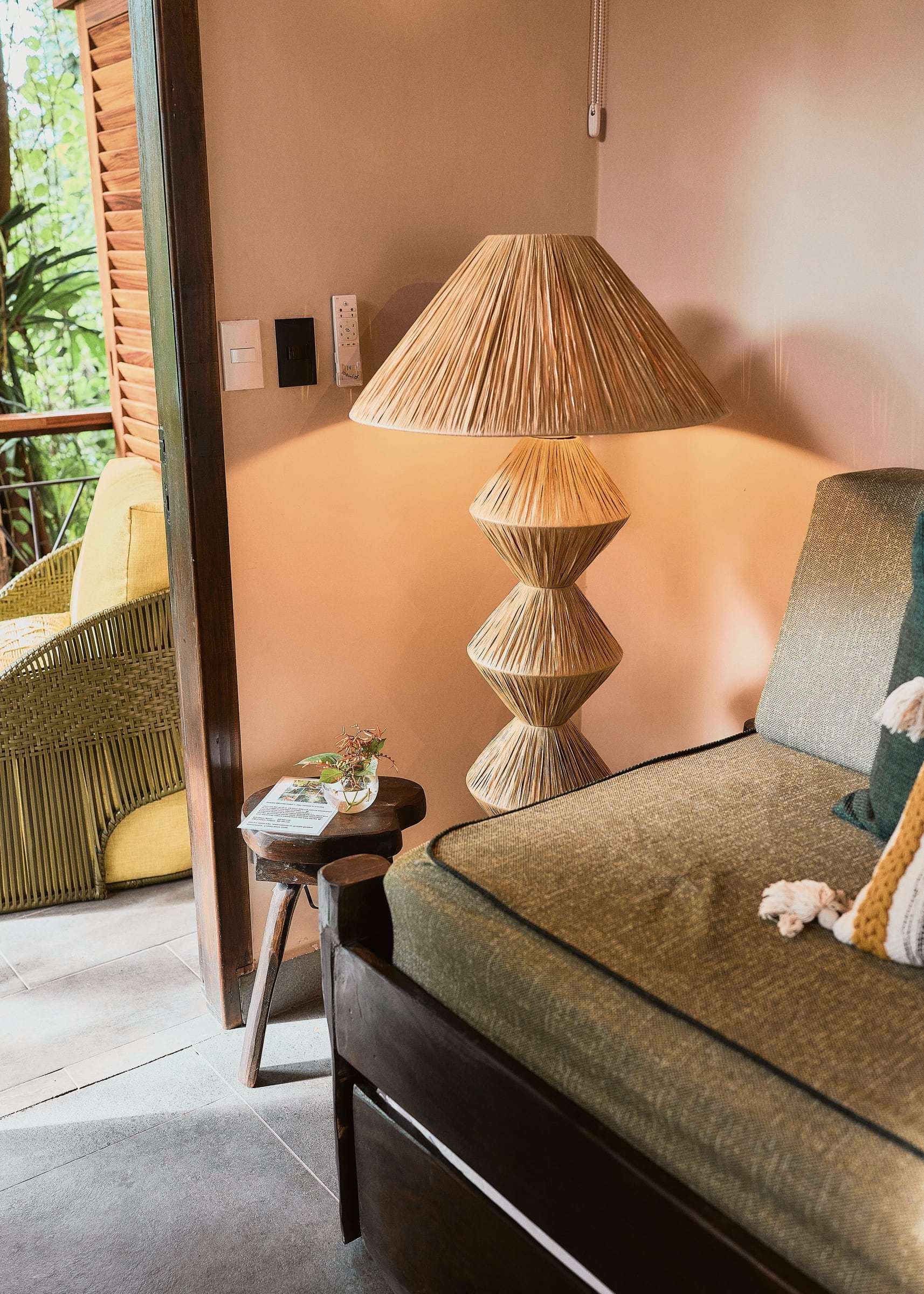 Jungle View Suite: raffia floor lamp beside a green daybed, with a yellow armchair on the terrace beyond the door