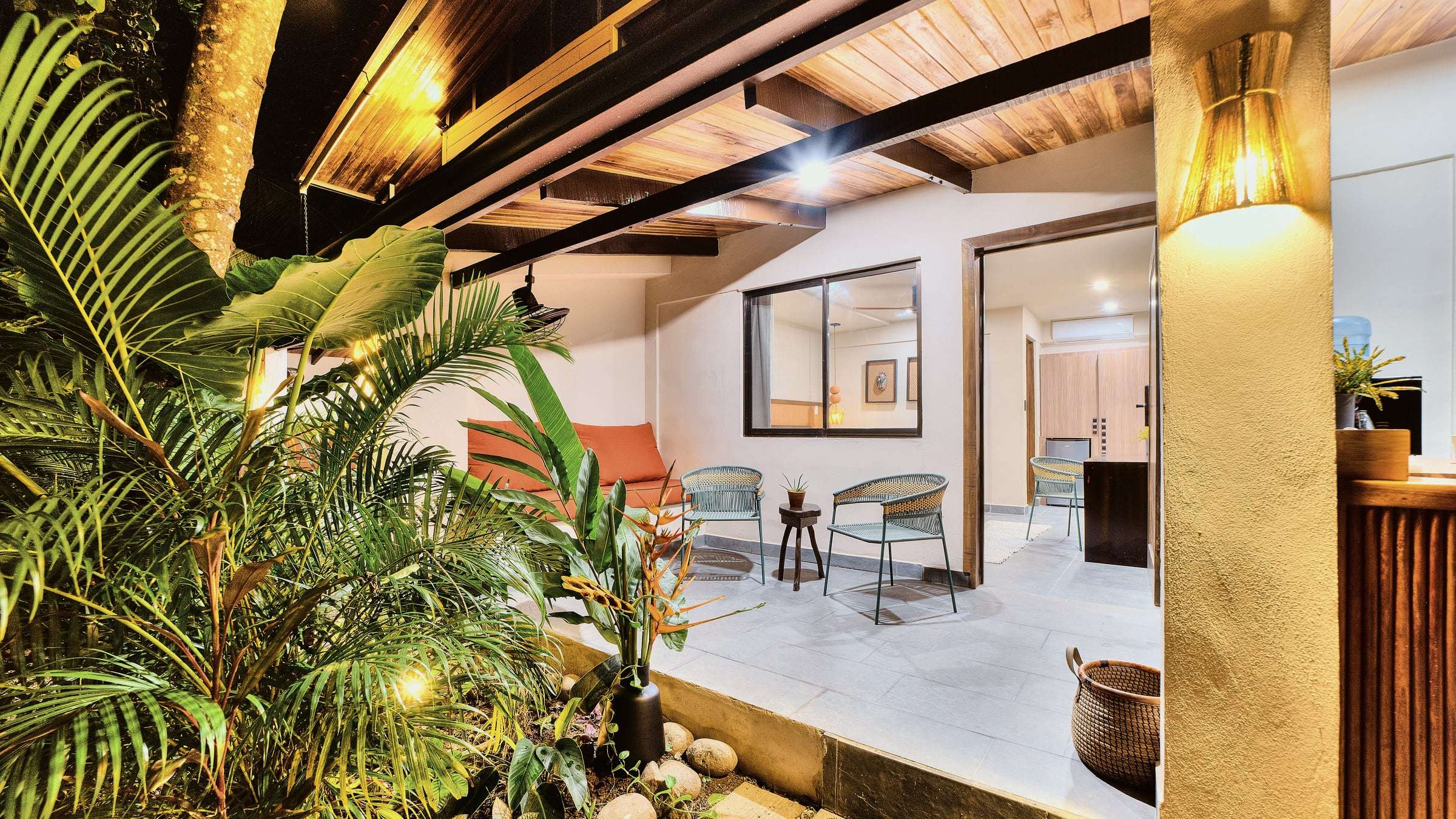 Ground-floor terrace at night with an orange sofa, woven chairs and lit palms, the door open into the room