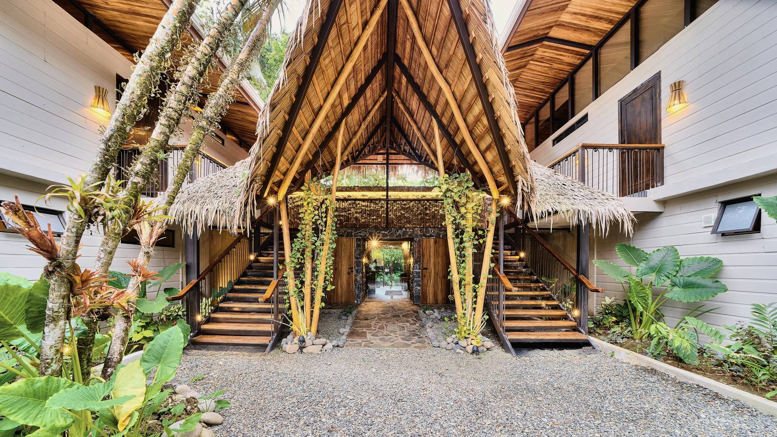 Palm-thatched entrance of Conga Boutique Hotel, with twin wooden stairs and a stone path leading through to the garden