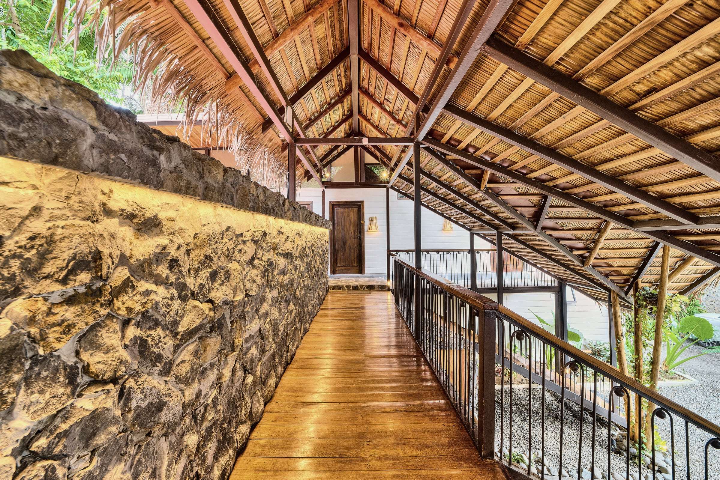 Upper walkway with a wooden floor, iron railing and palm-thatch roof, beside a warmly lit volcanic-stone wall