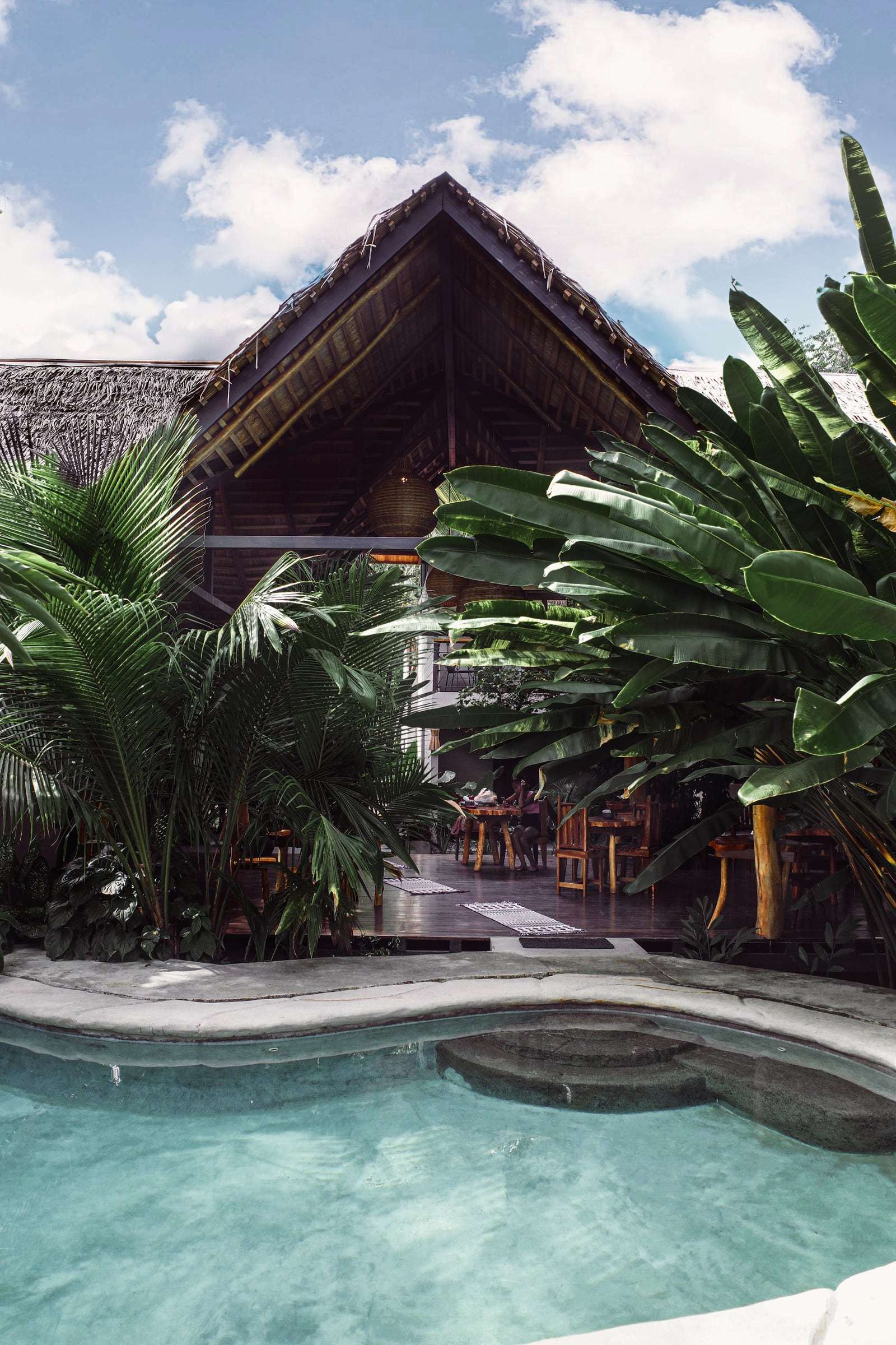 Turquoise pool in front of Conga's palm-thatched rancho, framed by banana leaves and palms under a cloudy sky
