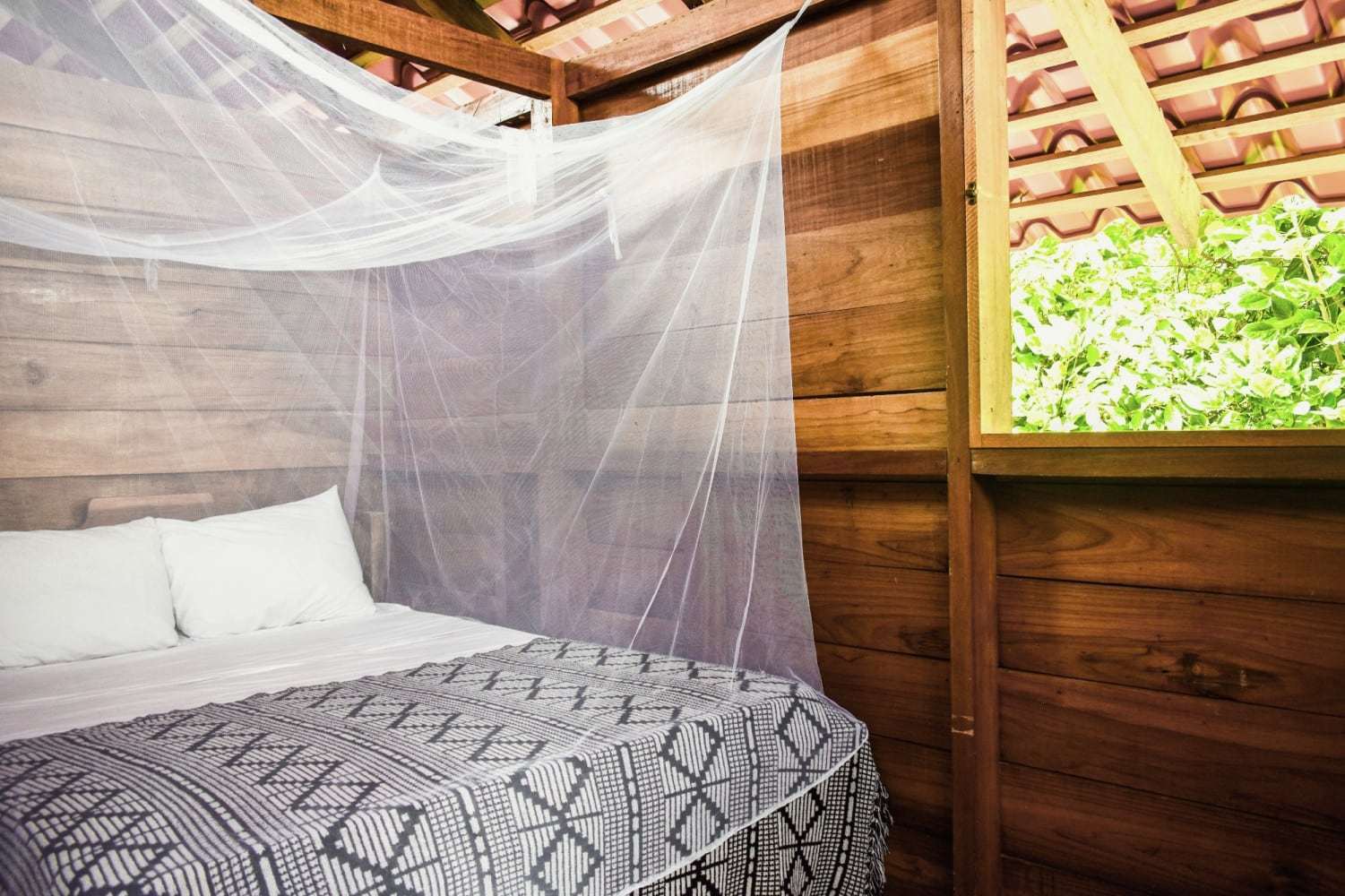 Garden Home bedroom: bed under a mosquito net, wood-plank walls and an open window onto the jungle