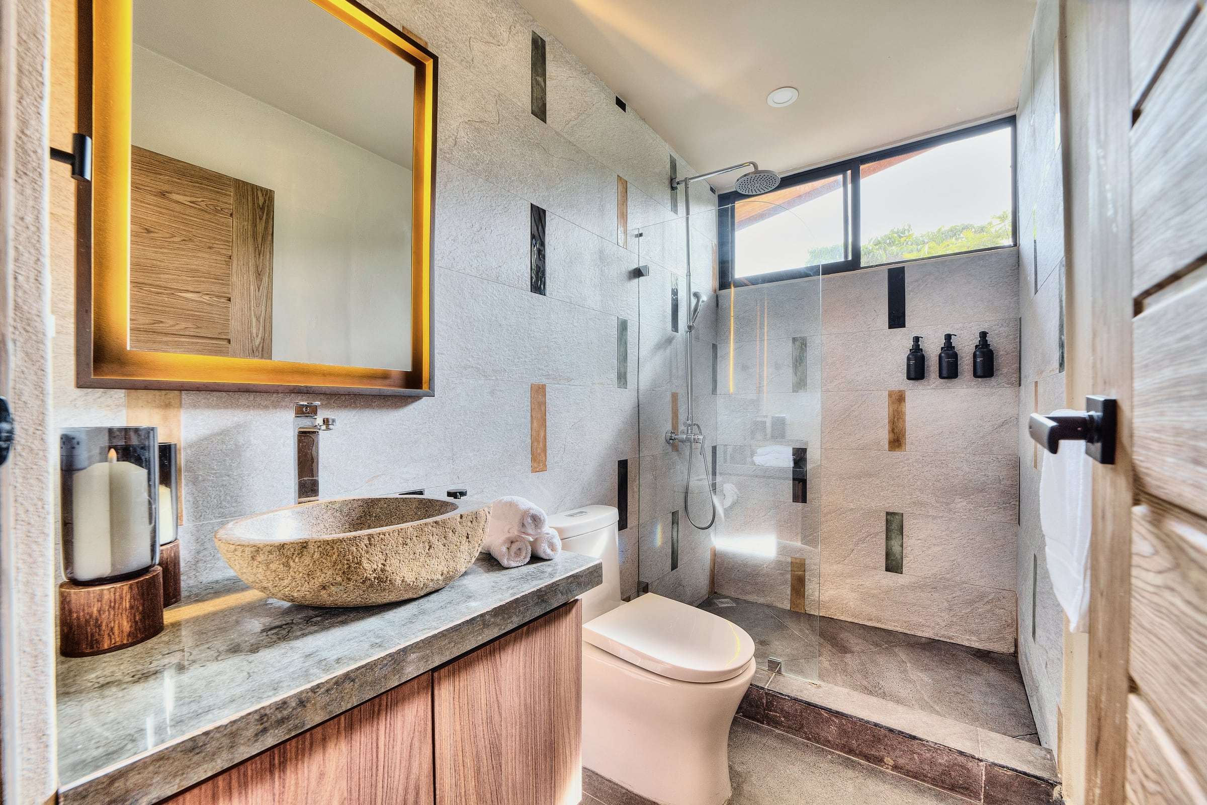 Jungle View Suite bathroom: river-stone vessel sink, backlit mirror and glass rain shower under a window to the trees