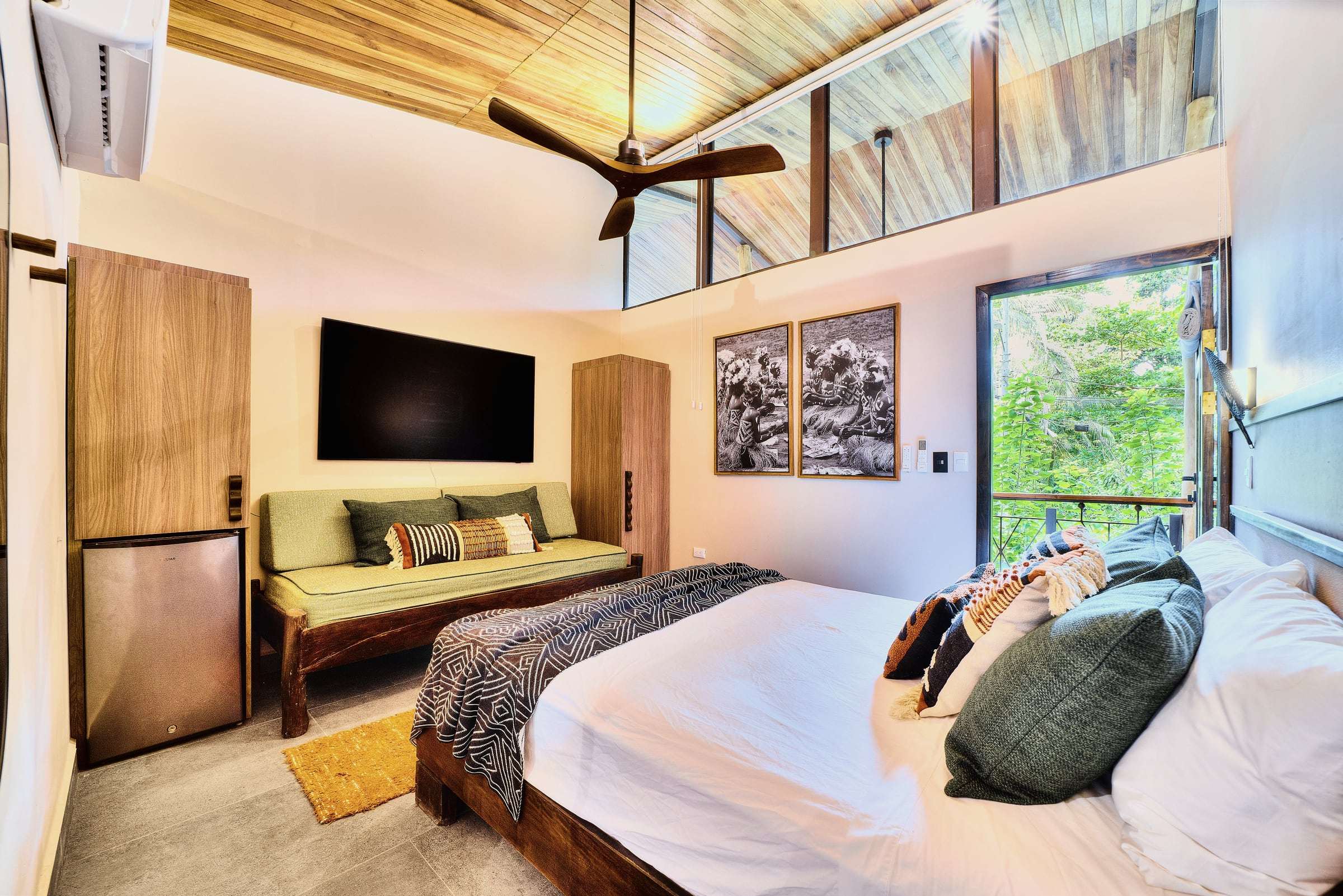 Terrace Suite: bed facing a green daybed and TV, black-and-white prints, high wood ceiling and a glass door to the trees