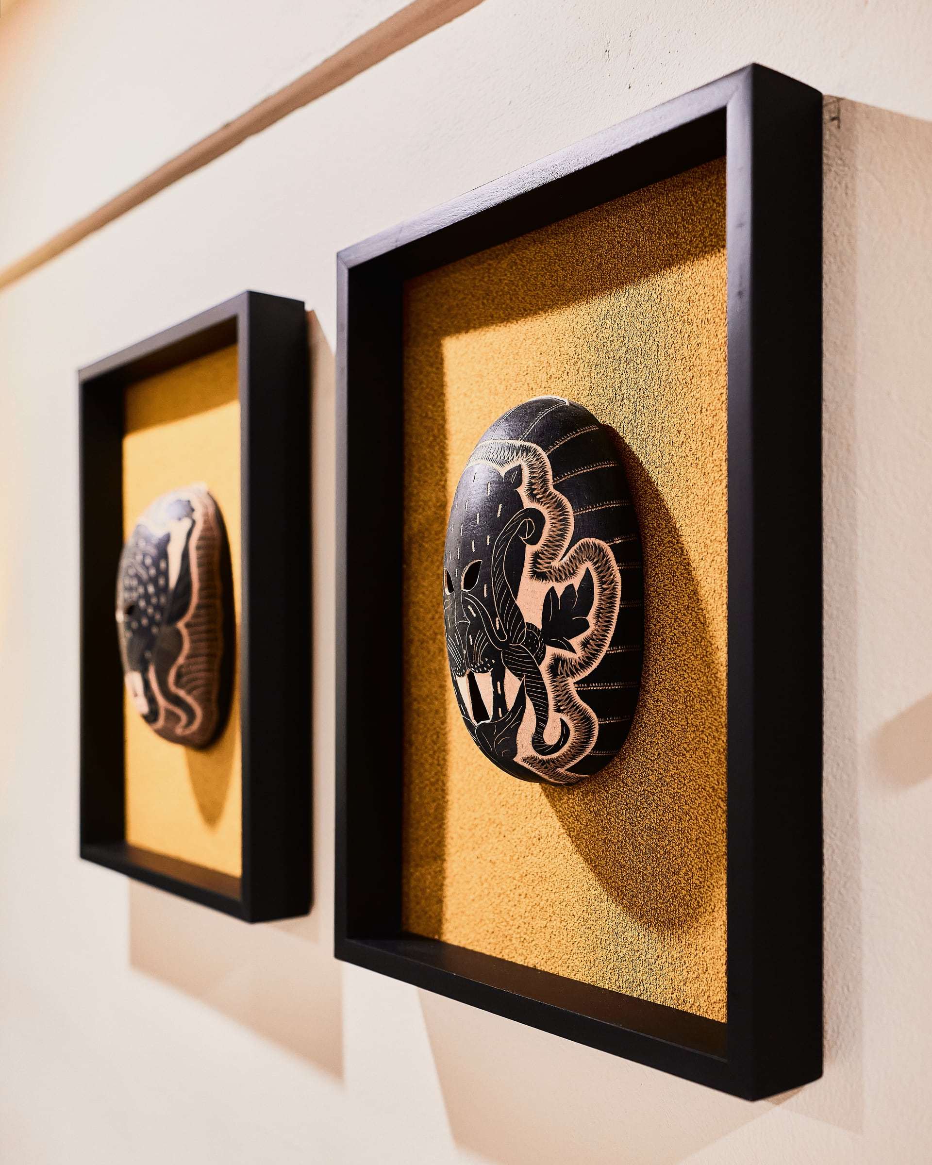 Hand-carved masks in black shadow-box frames on ochre backing, casting deep shadows on the wall